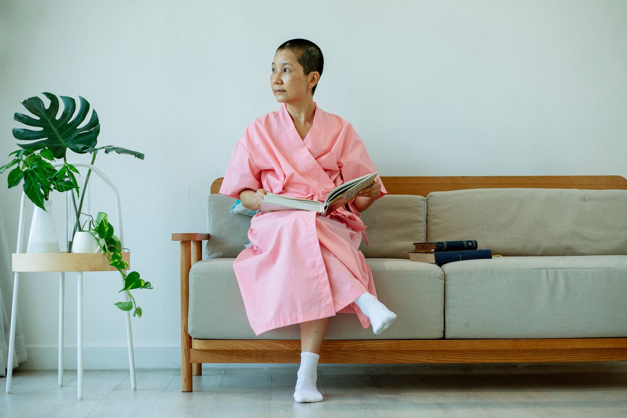 Thoughtful ethnic female in white socks relaxing with book on couch while looking away in hall of hospital